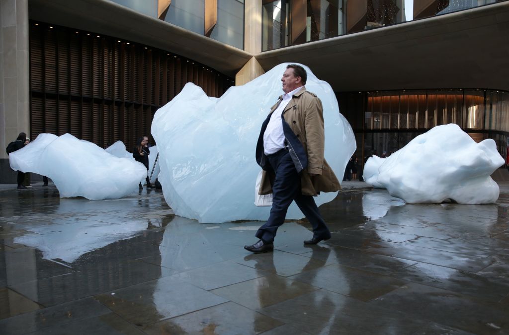 A man walks past a block of melting ice outside the Bloomberg building from an exhibit entitled 'Ice Watch' created by Icelandic-Danish artist artist Olafur Eliasson and leading Greenlandic geologist Minik Rosing outside Tate Modern in central London on December 11, 2018. - The blocks of ice were sourced from the waters of the Nuup Kangerlua fjord in Greenland. As the ice gradually thaws, members of the public have an opportunity to encounter the tangible effects of climate change by seeing and feeling the ice melt away. (Photo by Daniel LEAL-OLIVAS / AFP) / RESTRICTED TO EDITORIAL USE - MANDATORY MENTION OF THE ARTIST UPON PUBLICATION - TO ILLUSTRATE THE EVENT AS SPECIFIED IN THE CAPTION        (Photo credit should read DANIEL LEAL-OLIVAS/AFP via Getty Images)