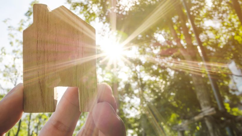 Hand holding wooden house with green forest background blurred and sun lighting