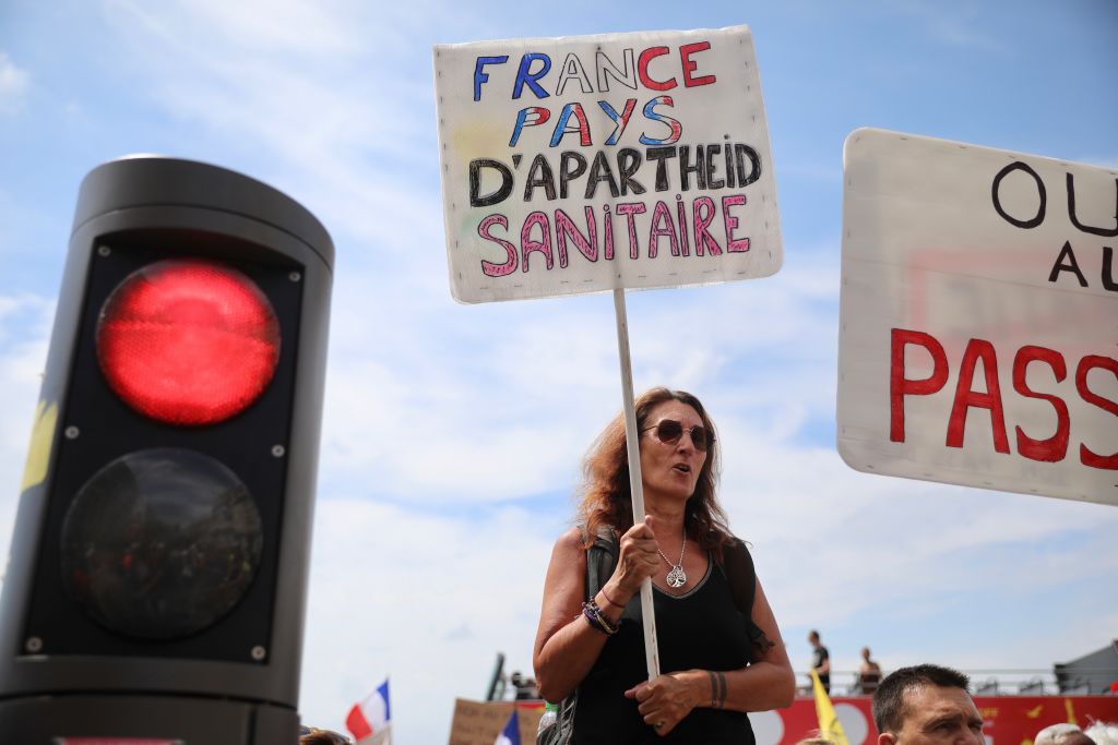 MARSEILLE, FRANCE - AUGUST 07: A demonstrator standing next to a red light holds up a placard that reads "France country of health apartheid" during a nationwide protest against France's new Covid-19 health pass on August 7, 2021 in Marseille, France.  The newly adopted Covid-19 health pass law drew crowds of demonstrators in more than 180 cities across France. The new health bill makes inoculation against Covid-19 compulsory for health and medical staff, firefighters, and certain army personnel. The health pass; either a negative test, vaccination certificate or certificate of recovery from Covid-19, entered into force in July in places of culture of more than 50 people and has been extended to include cafes, restaurants and inter-city trains and planes. (Photo by Roger Anis/Getty Images)