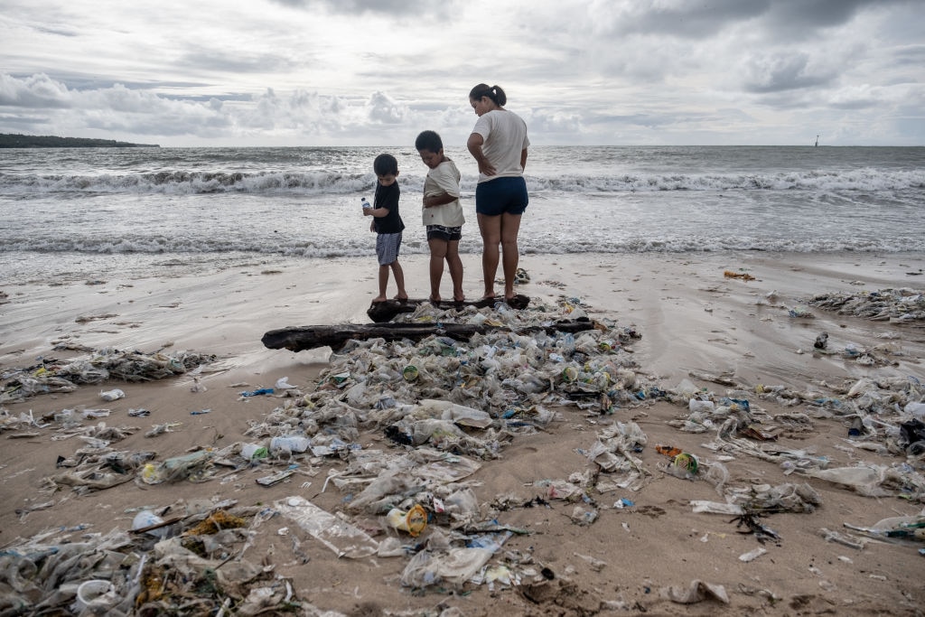A woman with two boys stand on some wood which is covered by plastic trash brought in by the strong waves during the northwest monsoon season at Jimbaran beach on January 27, 2021 in Jimbaran, Bali, Indonesia.(Photo by Agung Parameswara/Getty Images)