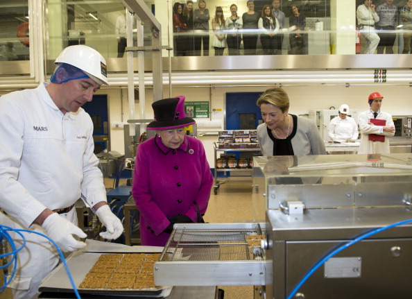 Queen Elizabeth II stands alongside Fiona Dawson, President of Mars Chocolate UK (R) as Paul Milligan (L) puts Mars bars into a machine to cover them with chocolate in the pilot plant, part of the research and development, during a visit to Mars Chocolate UK in Slough on April 5, 2013. (Photo by Adrian Dennis - WPA Pool/Getty Images)