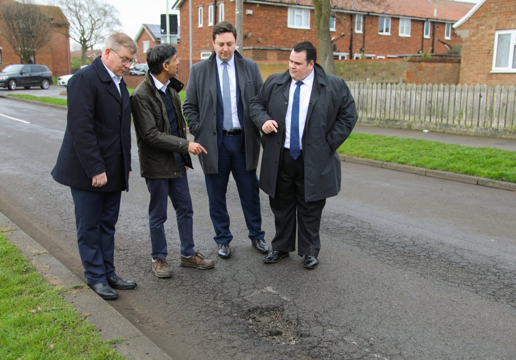 A gaggle of British decisionmakers stand around a pothole