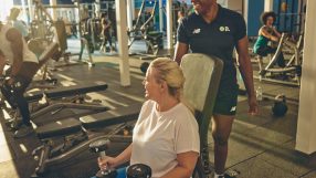 Woman lifting dumbbells with a trainer in a busy gym, promoting fitness and health