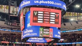 Plus500 branding on a large Jumbotron scoreboard at a US sports arena, displaying game stats.