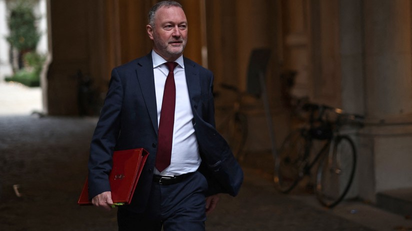 Steve Reed speaking at a press conference, addressing media with a serious expression, wearing a suit and tie.