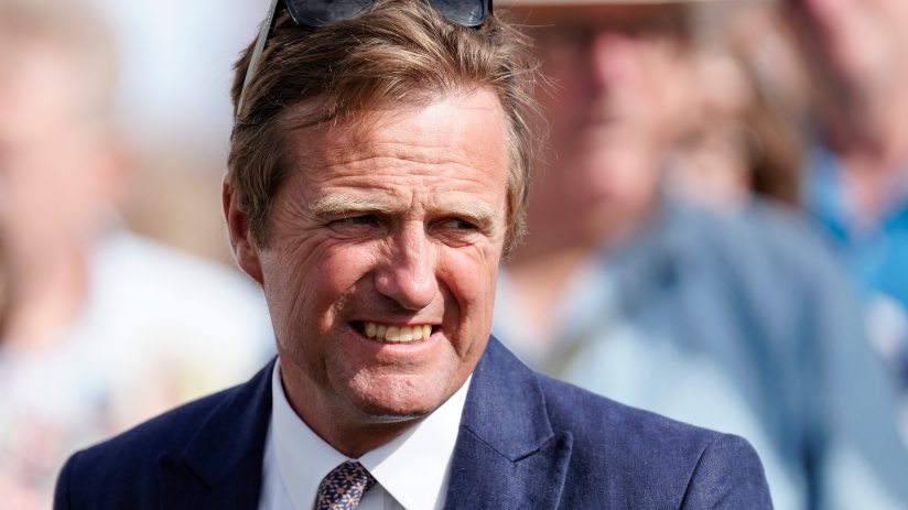 Ralph Beckett speaking at a press conference, wearing a suit, with microphones and reporters visible in the background