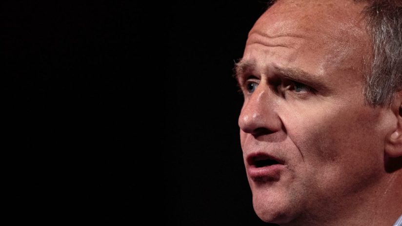 Sir Dave Lewis, former Tesco CEO, speaking at a business conference, wearing a suit and tie, emphasizing strategic insights