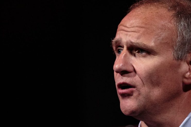 Sir Dave Lewis, former Tesco CEO, speaking at a business conference, wearing a suit and tie, emphasizing strategic insights