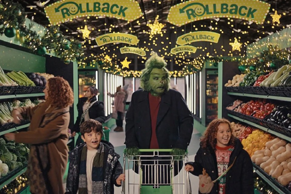 Grinch character wearing a Santa suit, holding a sack of presents, smiling mischievously in the Asda christmas advert