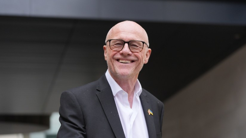 Breaking news event with a large, diverse crowd gathering in a city square under clear skies, capturing public interest.