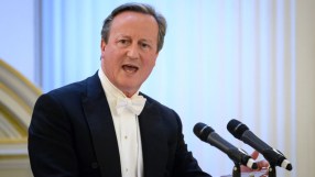 David Cameron smiling at a press conference podium, wearing a navy suit and blue tie, addressing the media in a formal set...