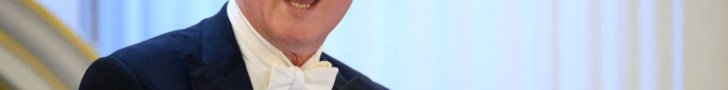 David Cameron smiling at a press conference podium, wearing a navy suit and blue tie, addressing the media in a formal set...