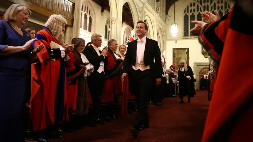 David Cameron speaking at a press conference, wearing a suit and tie, addressing the media with a serious expression.