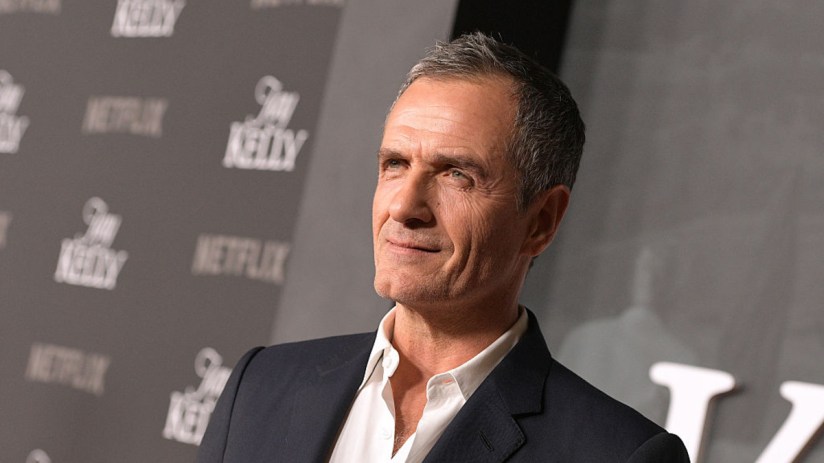 David Heyman speaking at a press conference, wearing a suit and tie, with a backdrop of industry event logos and banners.