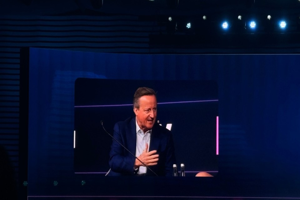 David Cameron delivering a speech as Prime Minister, standing at a podium with a UK flag backdrop, addressing economic pol...