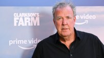 Jeremy Clarkson speaking at a press conference, wearing a suit, gesturing with his hands, addressing a crowd of journalists