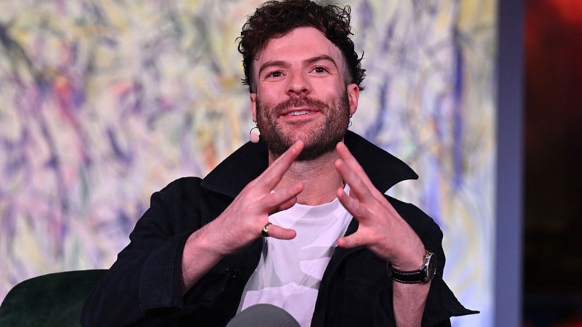 Jordan North in a studio setting, wearing headphones and speaking into a microphone during a radio broadcast.