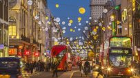 Busy Oxford Street with bustling shoppers, iconic red buses, and historic architecture under a clear blue sky.