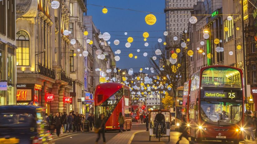 Busy Oxford Street with bustling shoppers, iconic red buses, and historic architecture under a clear blue sky.
