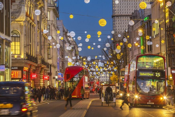 Busy Oxford Street with bustling shoppers, iconic red buses, and historic architecture under a clear blue sky.