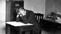 Pessimistic businessman sitting at a desk with a frown, surrounded by office documents and a computer, conveying workplace...