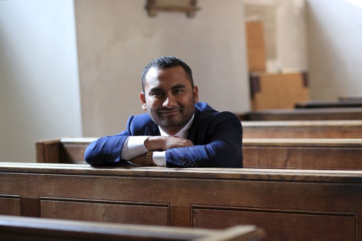Amol Rajan speaking at a press conference, wearing a suit and tie, addressing the media with microphones in front of him