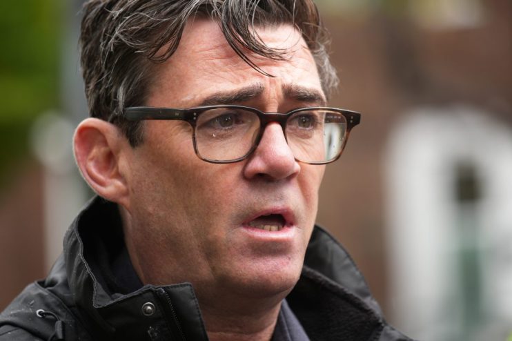 Andy Burnham speaking at a public event, addressing local community issues, wearing a suit and tie, engaged audience in ba...