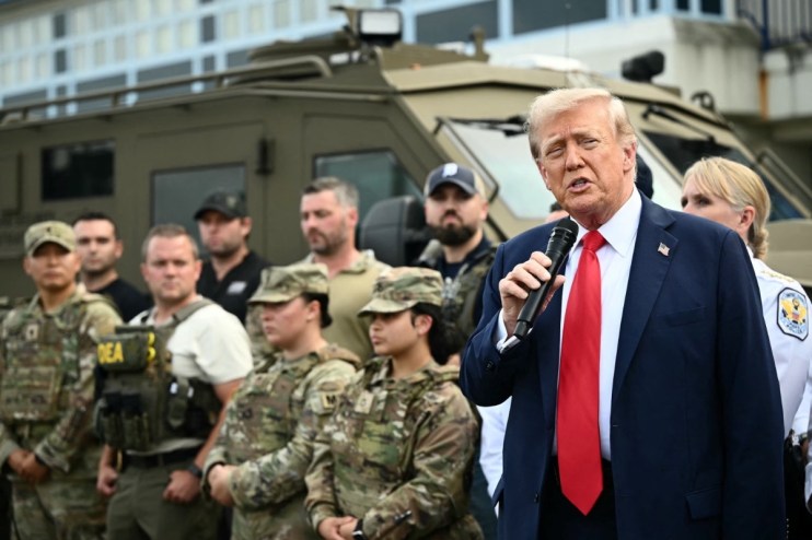 Donald Trump speaking at a press conference, addressing media questions, wearing a suit and tie, with American flags in ba...