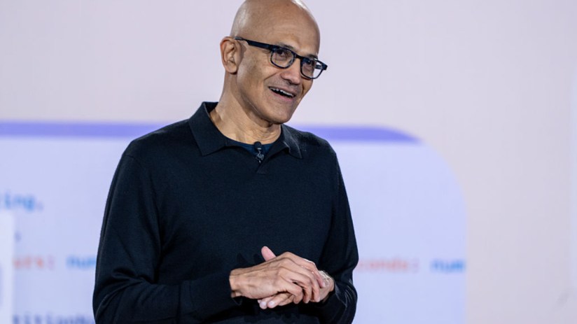 Satya Nadella, CEO of Microsoft, speaking at a tech conference, wearing a suit, with a backdrop of the Microsoft logo