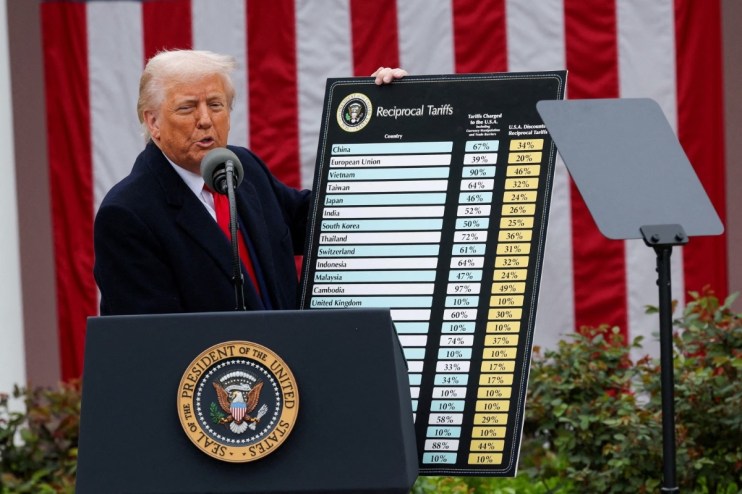 President Trump addressing the media during a press conference, wearing a navy suit and red tie, with the American flag in...