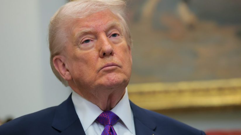 Donald Trump speaking at a press conference podium with an American flag backdrop, emphasizing political discourse