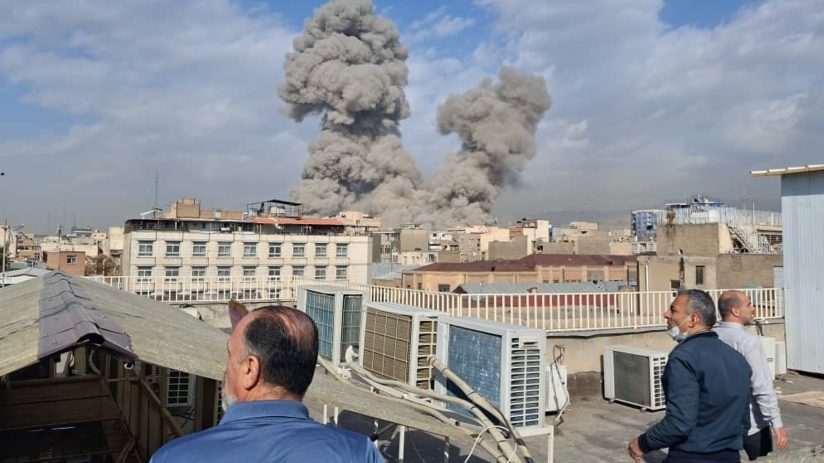Iran explosion aftermath showing smoke billowing from a building, emergency response teams on site, and debris scattered a...