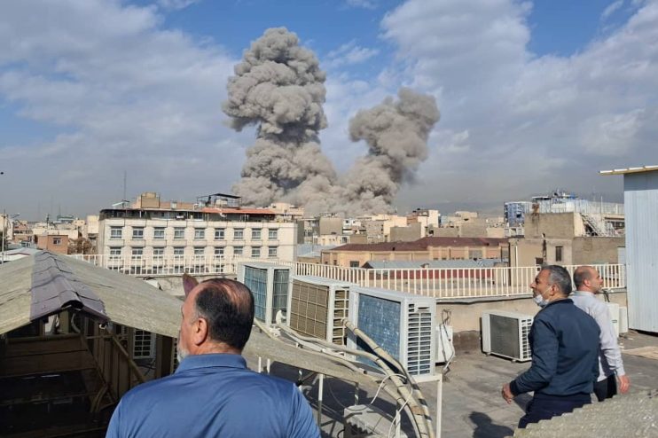 Iran explosion aftermath showing smoke billowing from a building, emergency response teams on site, and debris scattered a...