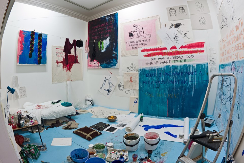 Tracey Emin standing in an art gallery, discussing her latest exhibition, surrounded by contemporary artworks and visitors