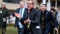 Warren Greatrex discussing racing strategies at a press conference, wearing a tailored suit and standing at a podium