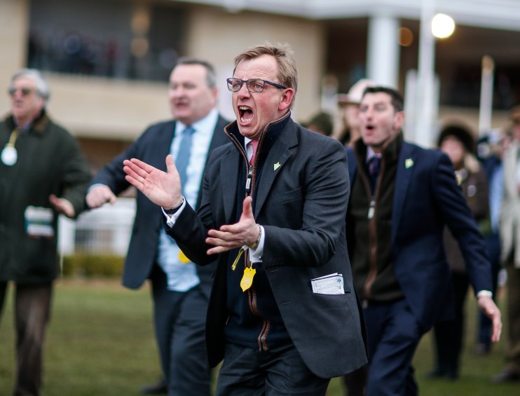 Warren Greatrex discussing racing strategies at a press conference, wearing a tailored suit and standing at a podium