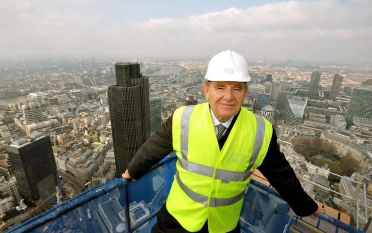 Gerald Ronson in a suit at a business conference, gesturing during a keynote speech, engaging with the audience.