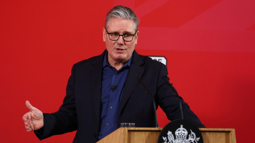 Prime Minister Keir Starmer addressing media at a public event, wearing a dark suit and tie, gesturing confidently