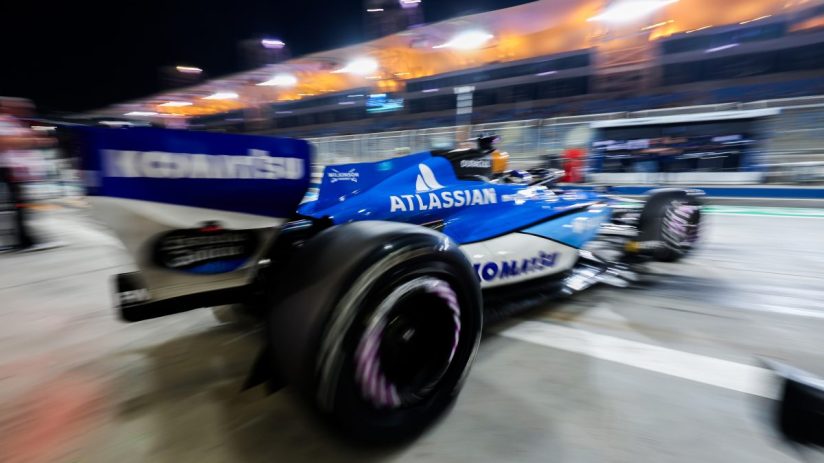 Alex Albon exits Formula 1 garage, wearing racing suit, surrounded by team members at busy pit lane during race weekend.
