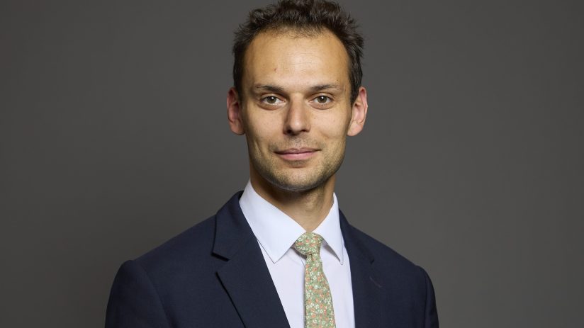 Josh Simons speaking at a business conference podium, wearing a suit, with a presentation screen in the background