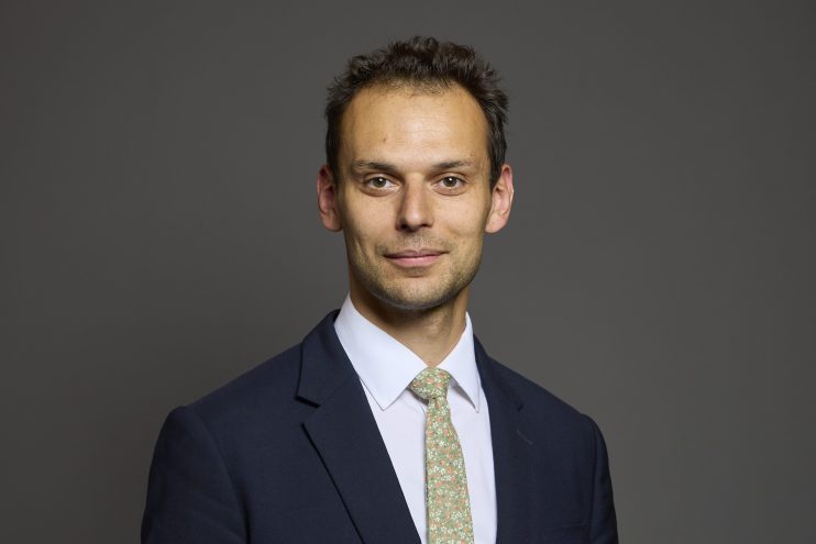 Josh Simons speaking at a business conference podium, wearing a suit, with a presentation screen in the background