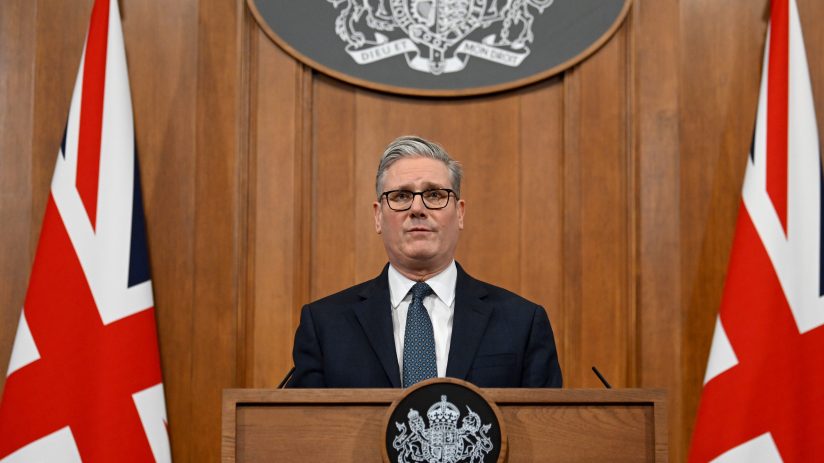 Keir Starmer discussing Iran policy at press conference podium with flags in the background