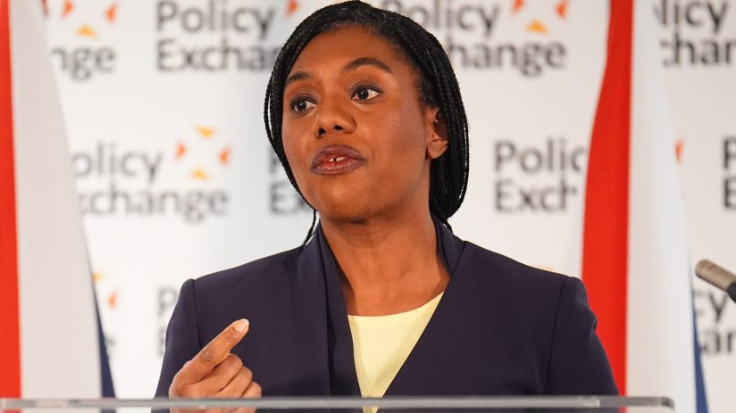 Kemi Badenoch speaking at a podium during a press conference, wearing a navy blazer and addressing the audience confidently