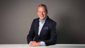 Michael Hayman in a professional headshot, wearing business attire, smiling confidently, suitable for a news profile feature.