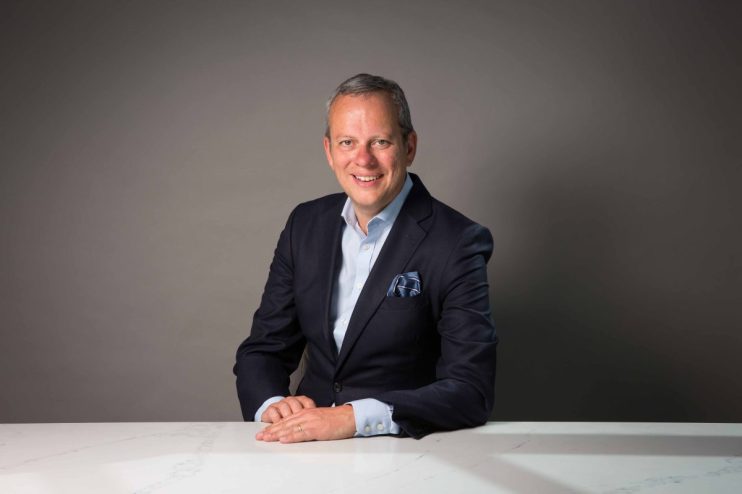 Michael Hayman in a professional headshot, wearing business attire, smiling confidently, suitable for a news profile feature.