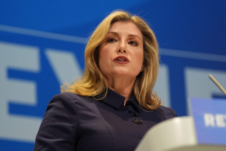 Penny Mordaunt delivering a speech at a podium, dressed in business attire, addressing a professional audience