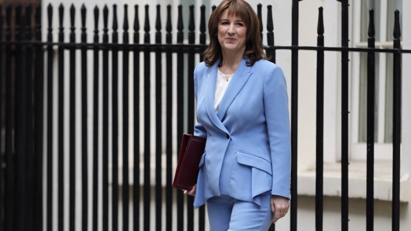 Rachel Reeves delivering spring statement at podium with financial charts in background, addressing economic policies.