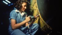 Rosie the Riveter-inspired worker in a factory setting, wearing a blue jumpsuit and red bandana, using a riveting tool.
