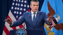 Pete Hegseth delivering a speech at a news event, wearing a suit and tie, with a focused expression and lively audience ba...
