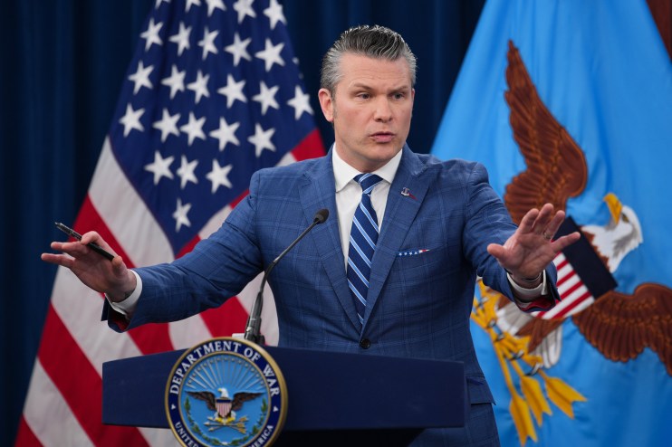 Pete Hegseth delivering a speech at a news event, wearing a suit and tie, with a focused expression and lively audience ba...